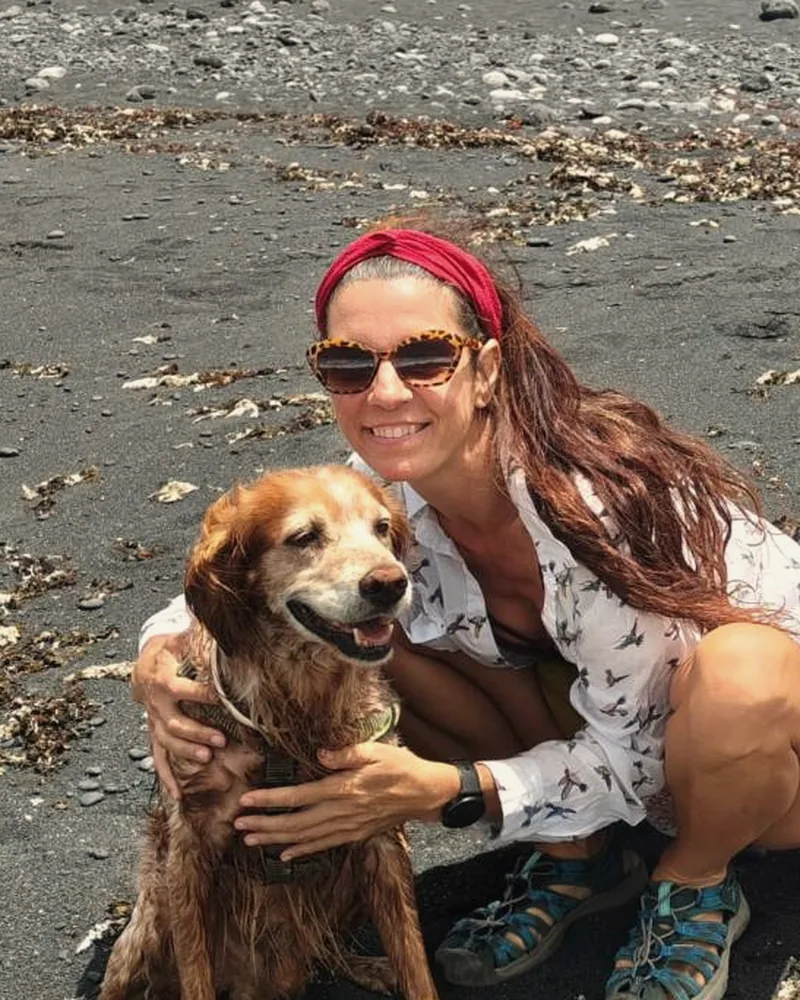 Retrato de Elena Rodríguez Martín, sonriendo y agachada en una playa de arena negra, abrazada a su perro.