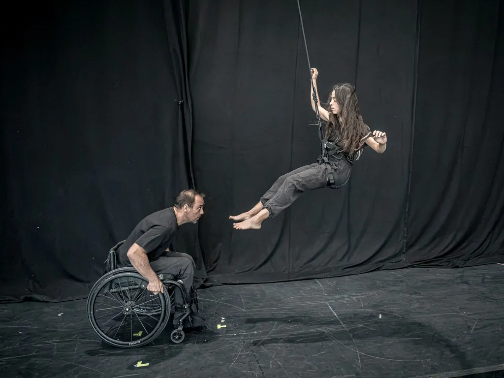 Durante una residencia de creación, una artista suspendida de una cuerda se balancea hacia un compañero en silla de ruedas sobre un escenario negro.